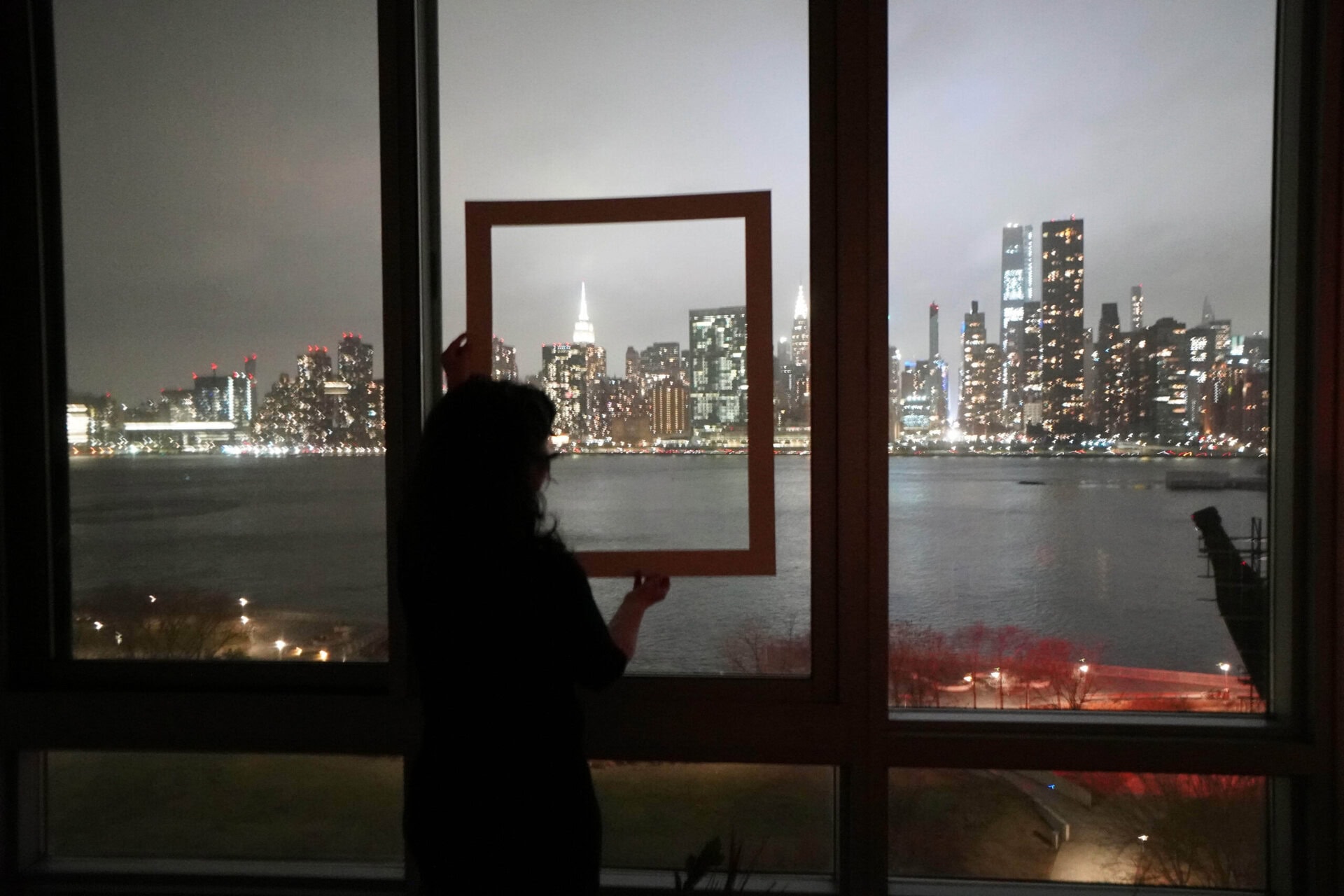 Night view of the New York City skyline seen from Long Island City, with illuminated buildings and reflections on the water.