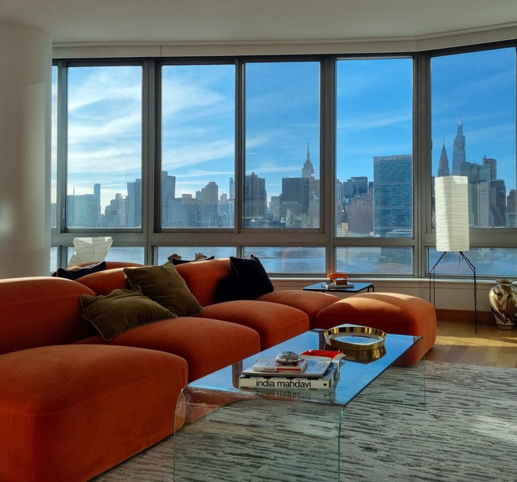 Luxury modern living room with floor-to-ceiling windows, natural sunlight, and a burnt orange modular sofa overlooking the New York City skyline.