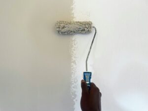 A hand applying a light beige paint sample onto a white wall using a paint roller.